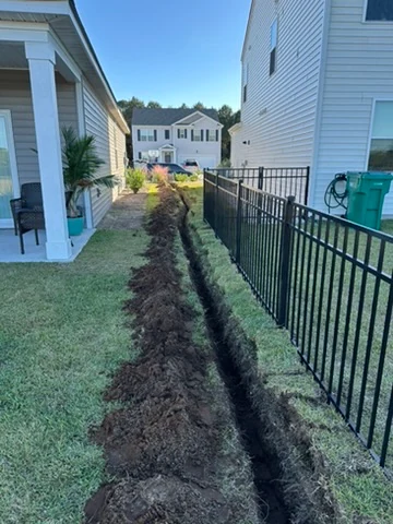 Open trench running beside a home and a black metal fence during outdoor work