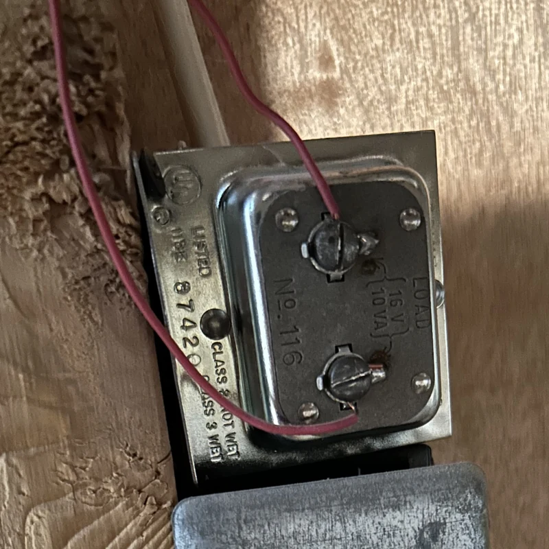 Metal doorbell transformer with two low-voltage screw terminals and red wires above an electrical box.